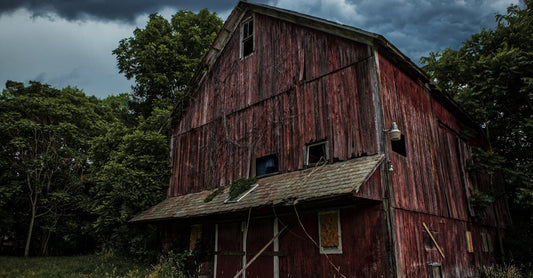 A rustic wooden barn in a rural landscape under dark storm clouds, surrounded by trees.