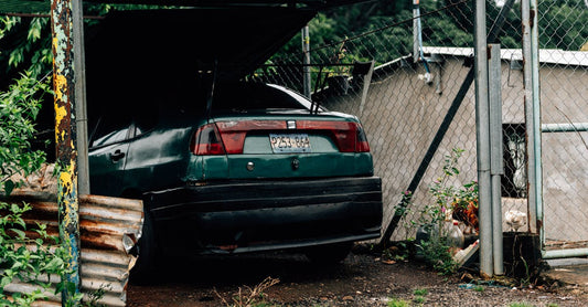 A green car parked under a corrugated metal roof by a chain-link fence, surrounded by greenery.