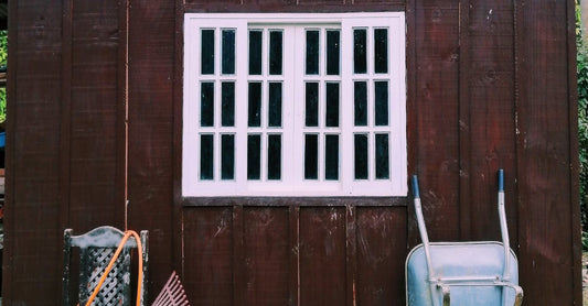 Simple wooden shed exterior showing tools like rake and wheelbarrow indicating gardening.