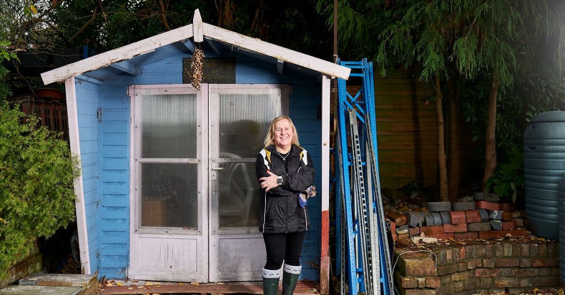 A happy woman stands in front of a blue shed, surrounded by garden equipment.